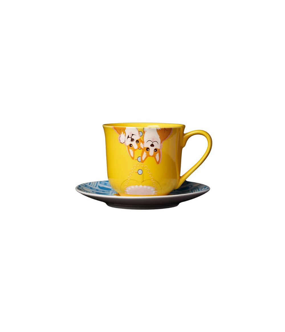 una vista ravvicinata della Tazza con Piattino The Queen, con la tazza gialla decorata con i corgi e il piattino blu con motivi iconici della monarchia britannica.