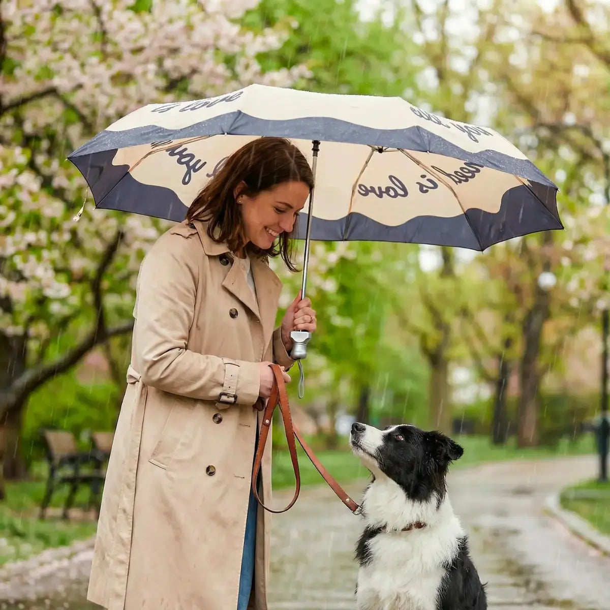 Donna con cane bianco e nero sotto all'ombrello tascabile doppio nero e crema con la scritta nera all you need is love. Sfondo di un parco con pioggia