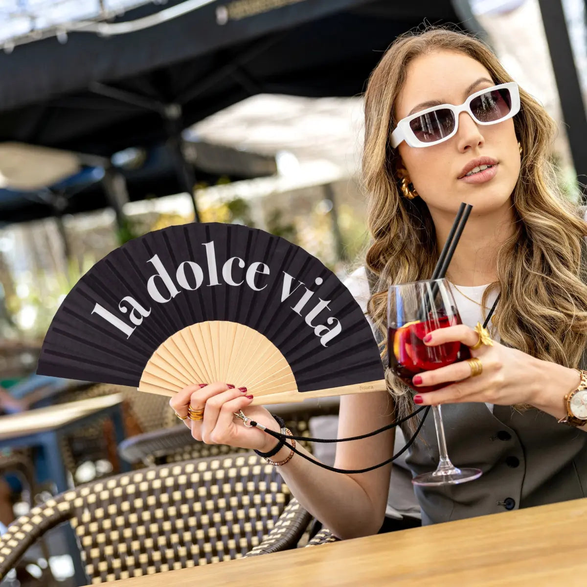 Donna al bar con appeso al collo il ventaglio la dolce vita nero, laccetto nero. Sfondo tavoli,sedie.