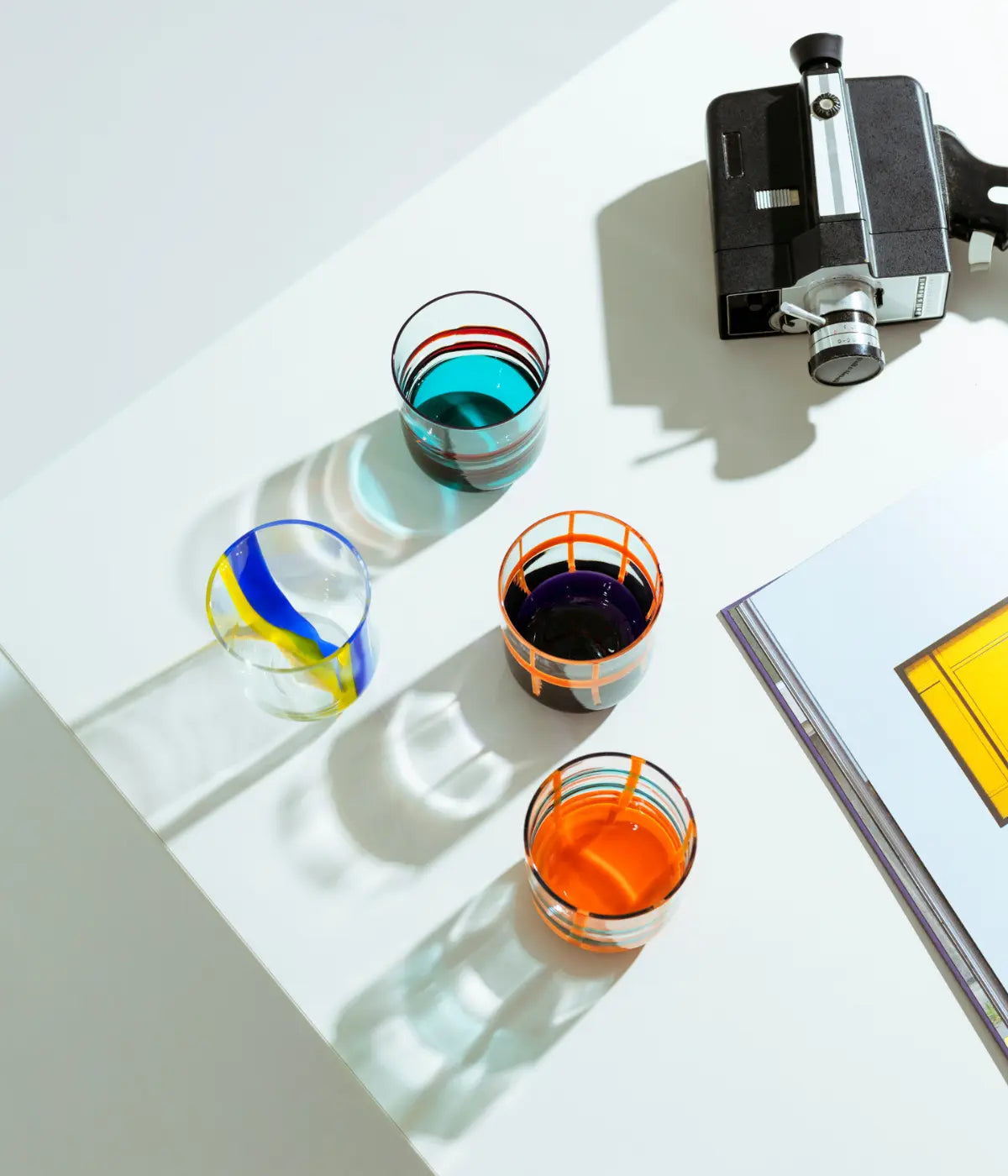 Quattro bicchieri con fondo colorato e righe. Appoggiati su tavolo bianco con cinepresa e libro