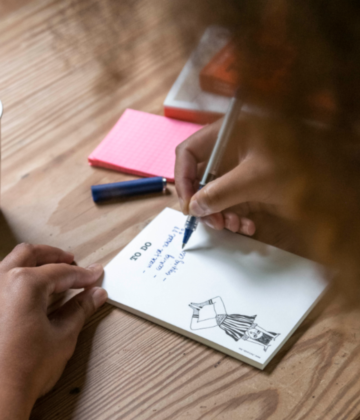 Persona che scrive su Blocknote bianco con ragazza a testa in giù e scritta To Do. Sfondo legno.