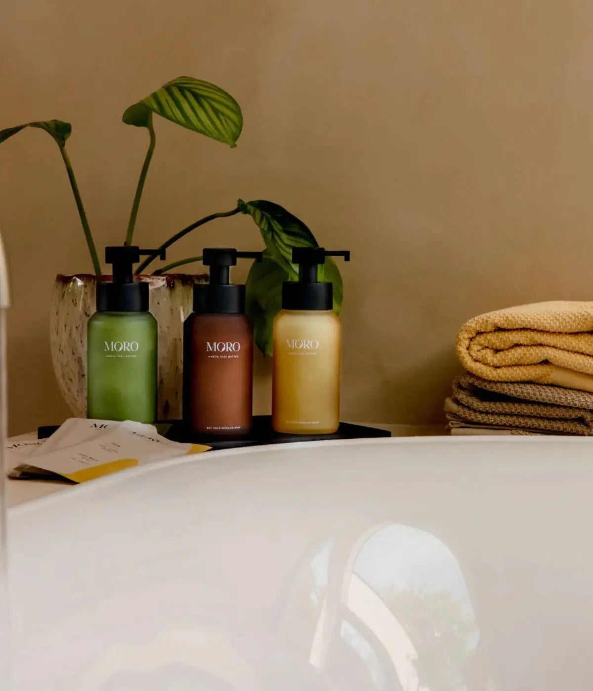 Bordo vasca bianca con i tre dispenser, verde, giallo e marrone su vassoio nero. Sfondo bagno