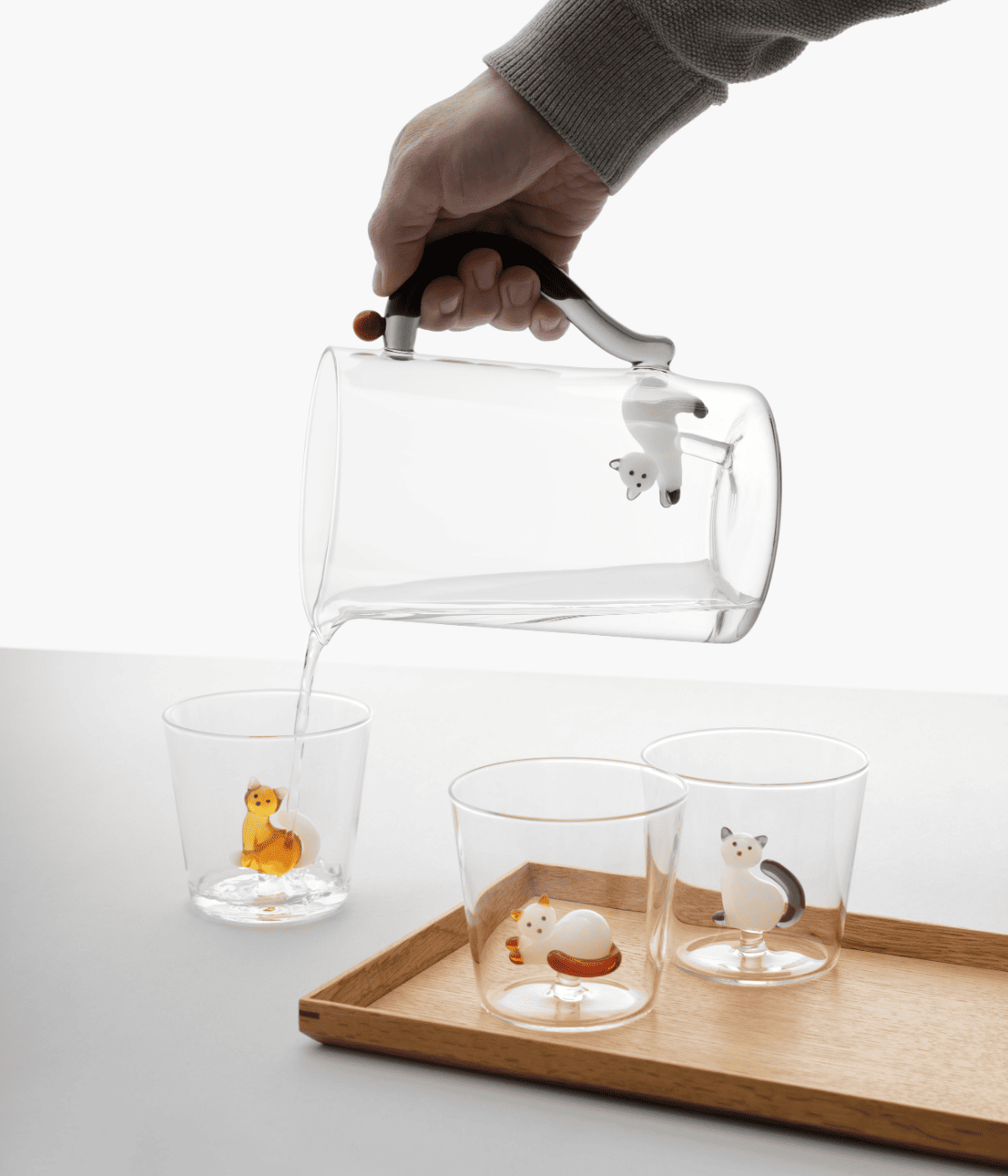 Brocca in vetro trasparente con gattino bianco e grigio. Bicchieri con gatto all'interno. Sfondo bianco con vassoio marrone.
