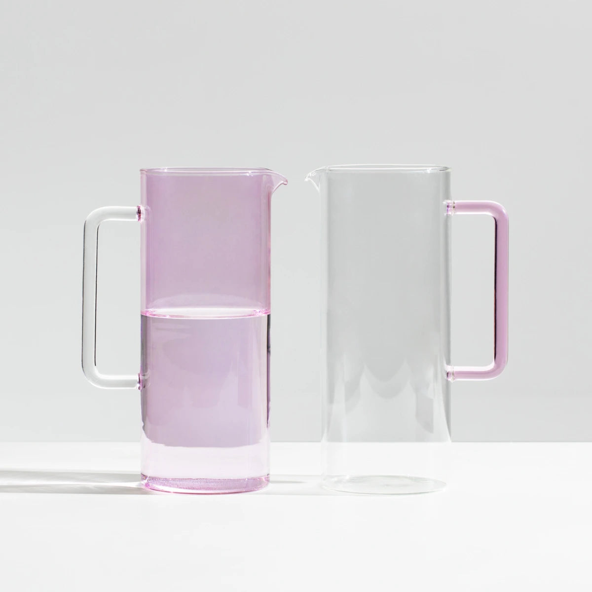 Due brocche in vetro una rosa, una trasparente appoggiate su tavolo bianco.
Sfondo muro bianco