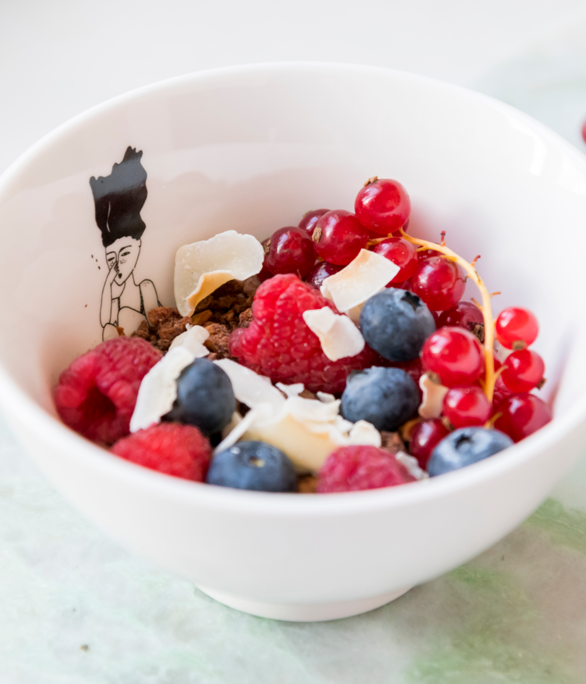 Ciotola bianca con cereali, lamponi, mirtilli. Disegno ragazza che si tuffa. Sfondo tavolo verde.