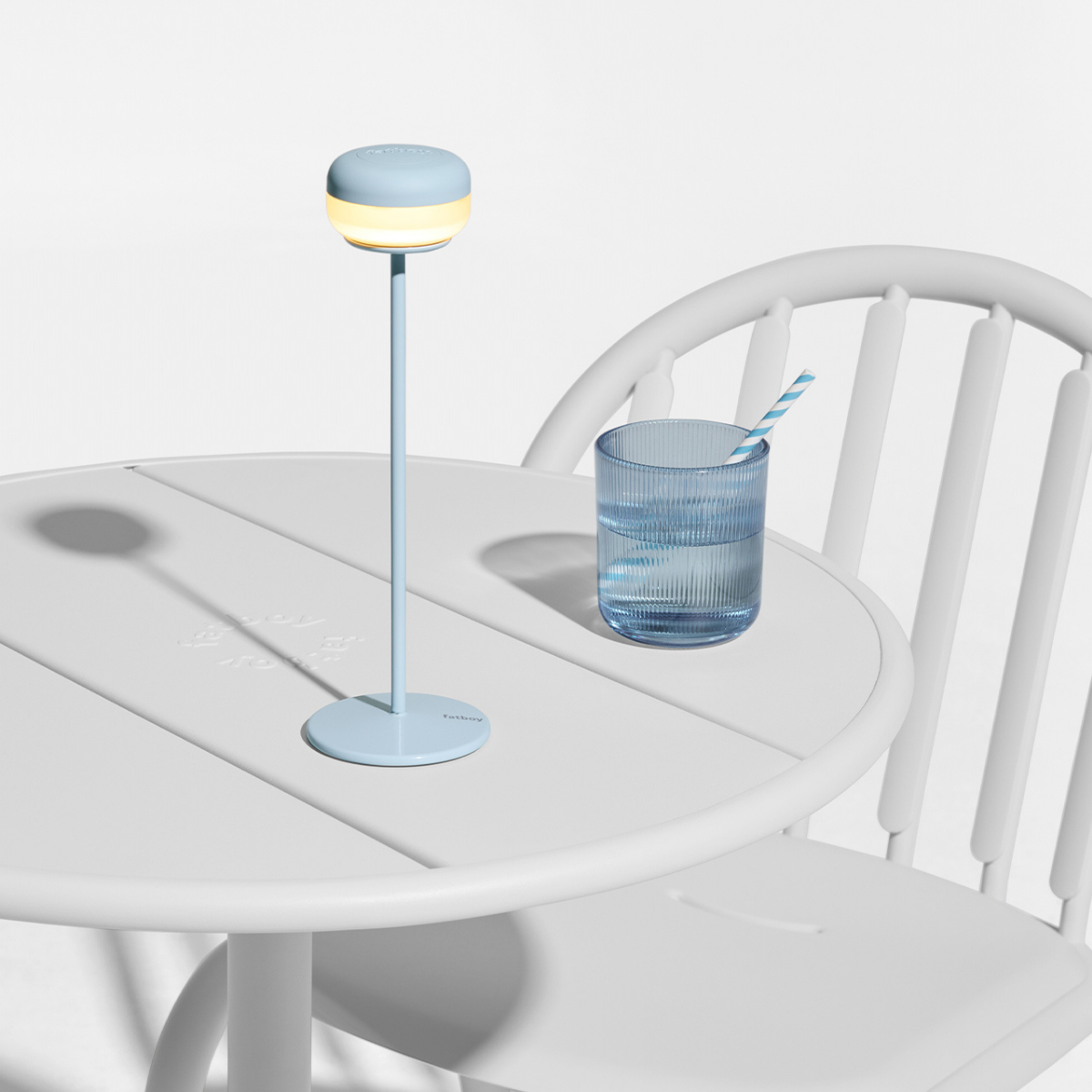 Lampada da tavolo azzurra appoggiata su tavolo bianco con vicino bicchiere d' acqua. Sfondo bianco