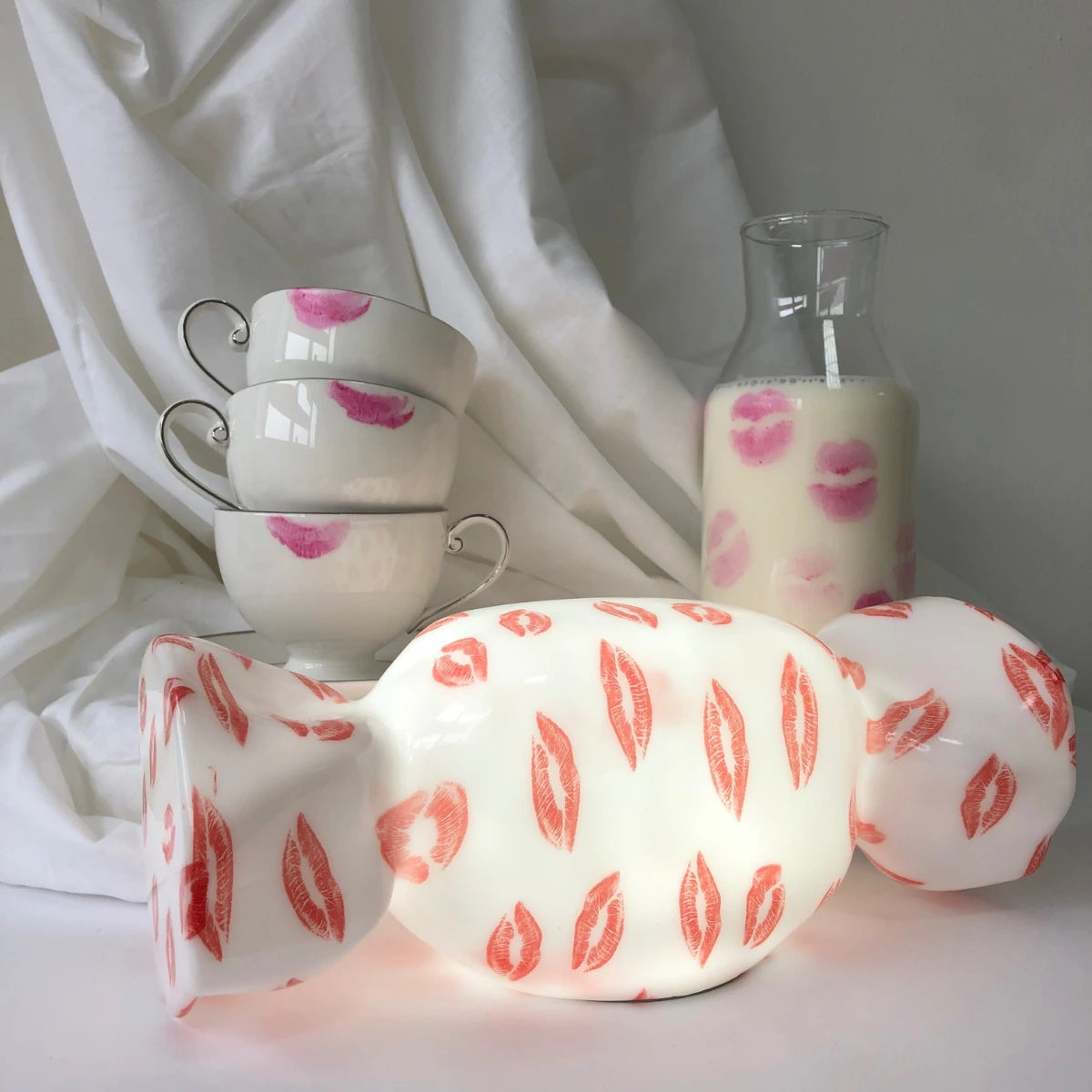 Lampada da tavolo a forma di caramella bianca con labbra rosse. Sfondo lenzuolo, tazza e brocca.