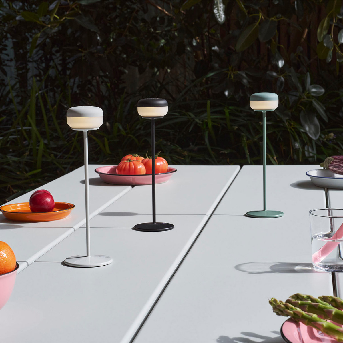 Tre lampade da tavolo beige, nero, verde salvia, appoggiate su tavolo bianco. Sfondo giardino