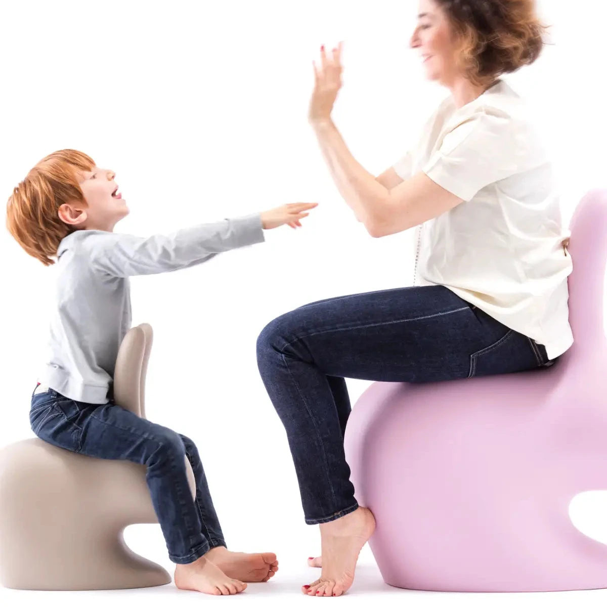 Una donna e un bimbo a cavalcioni su sedie a forma di coniglio una rosa e una grigia. Sfondo bianco