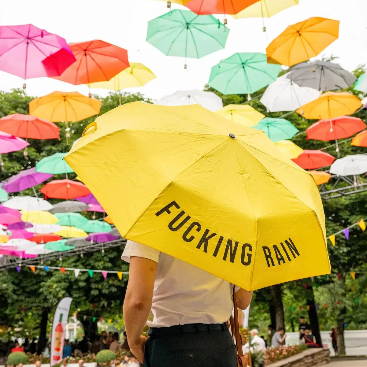 Persona che tiene ombrello giallo con scritta Fucking Rain. Ombrelli colorati sospesi.