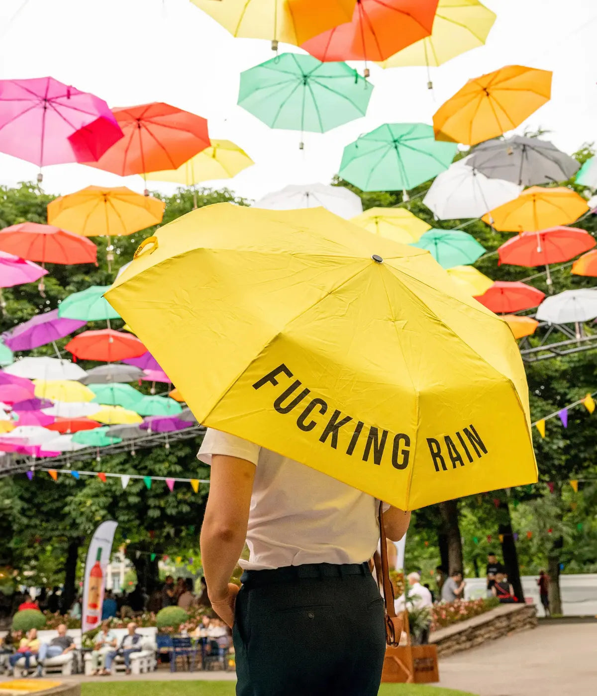 Persona che tiene ombrello giallo con scritta Fucking Rain. Ombrelli colorati sospesi.