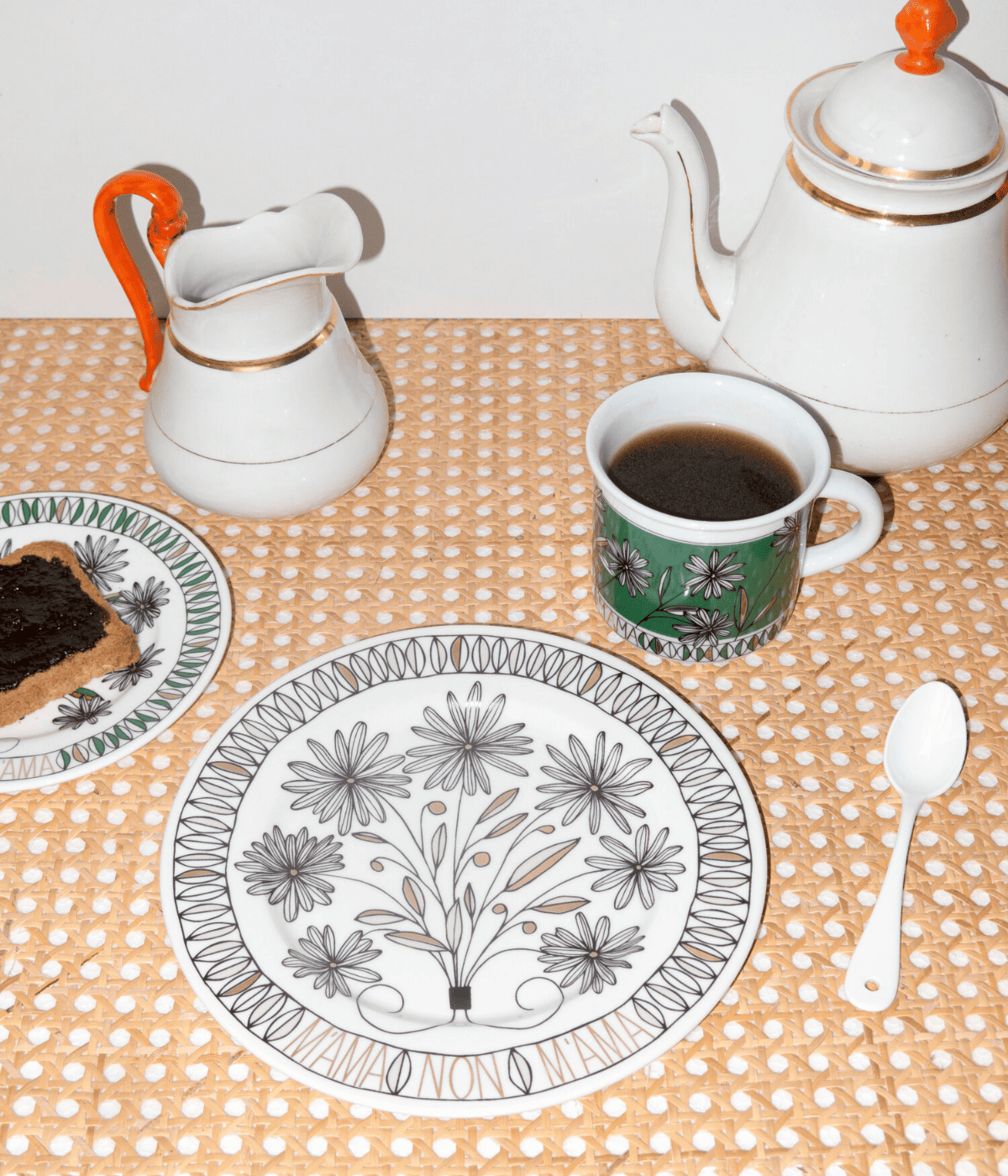 Piatto bianco con scritta oro. Decorazione floreale nero e oro. Sfondo tavolo con tazza e piattino abbinato. Teiera, brocca e cucchiaio bianco.