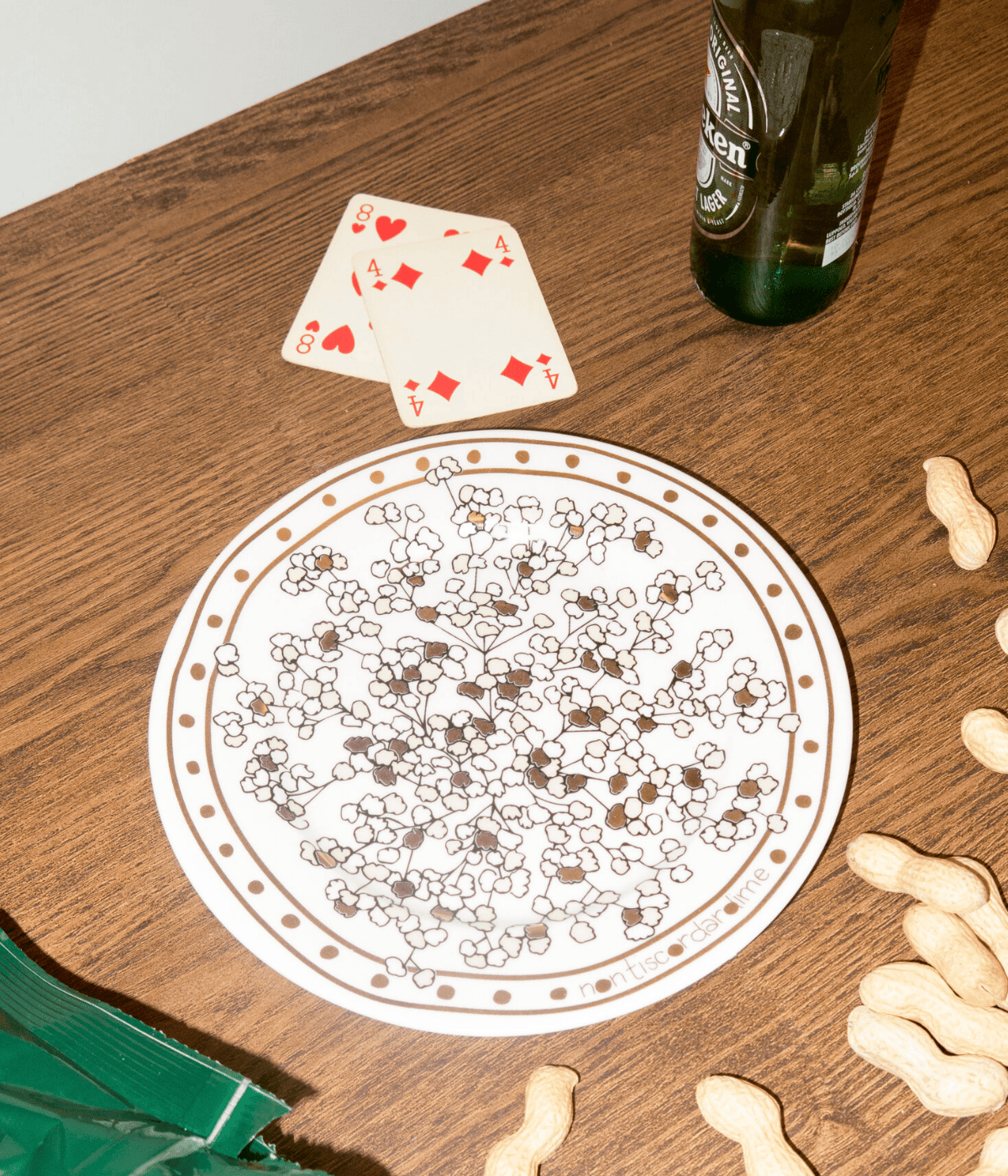 Piatto bianco con scritta oro e decorazioni floreali nero e oro. Sfondo tavolo marrone, carte, birra e arachidi.