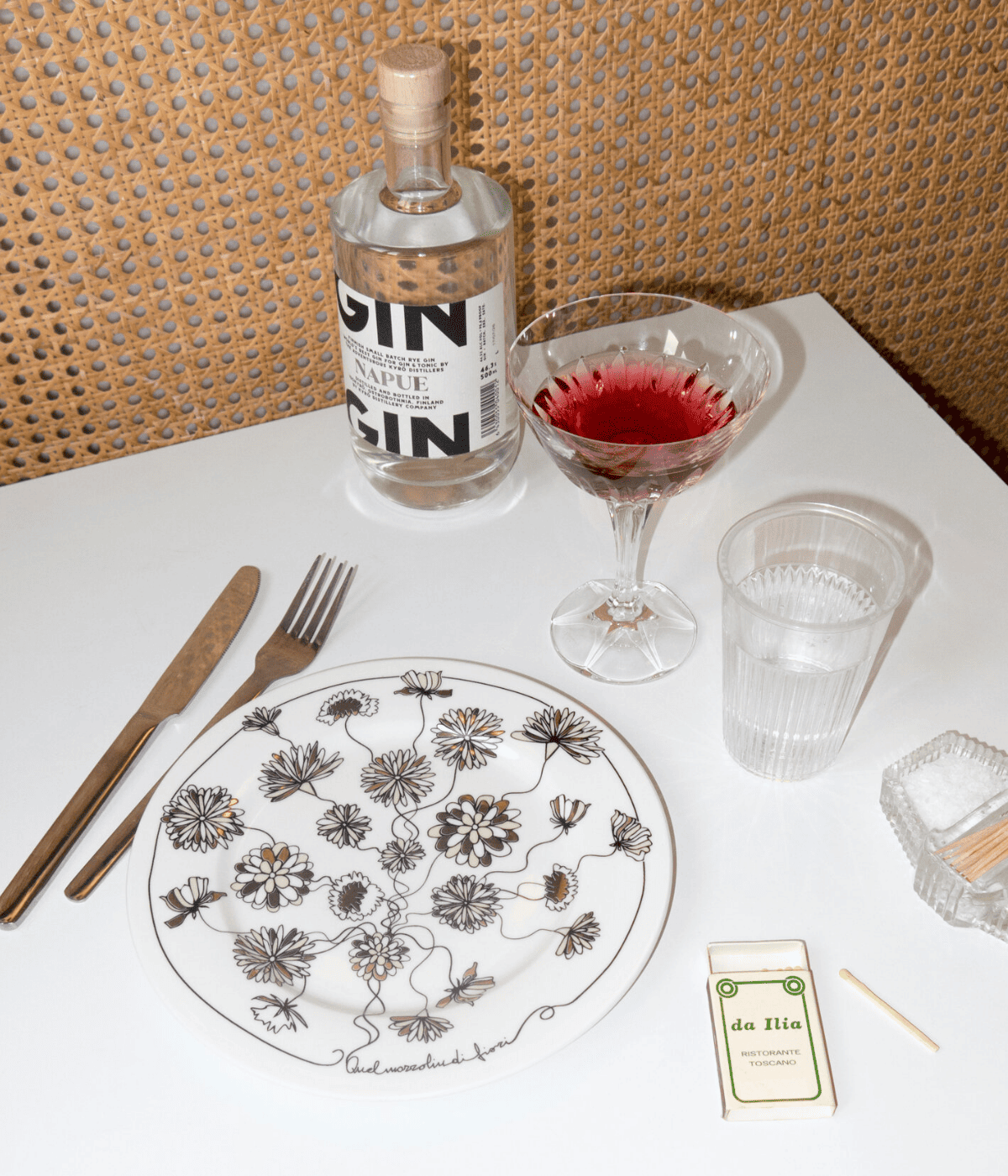 Piatto bianco con scritta nera e decorazioni floreali nero e oro. Sfondo tavolo bianco con poste, bottiglia di Gin e bicchieri.