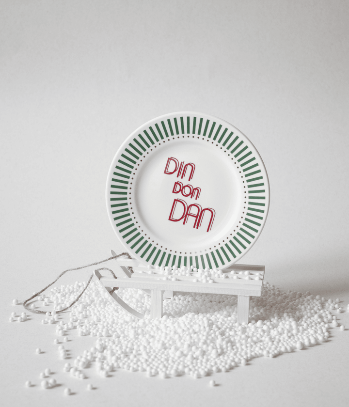 Piatto con scritta Din Don Dan e decorazioni verdi con puntini oro, appoggiato su slitta bianca e polistirolo. Sfondo bianco.
