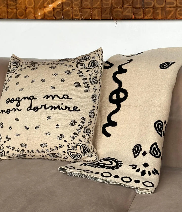 Un divano con sopra coperta e cuscino beige abbinati. Scritta sogna ma non dormire. 
