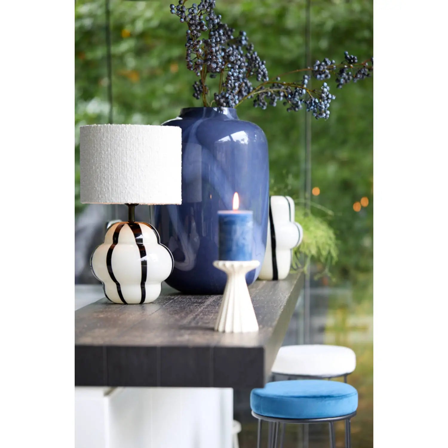 Un portacandele crema con candela blu sopra appoggiato su tavolo con lampade e vasi. Sfondo giardino