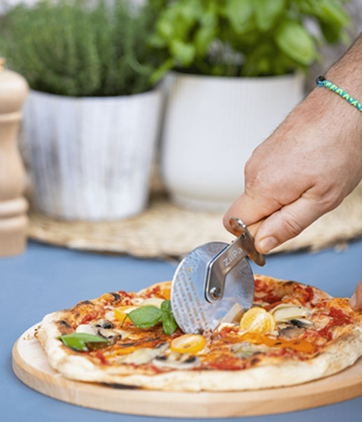 Mano che impugna rotella grigia, con scritta Ziipa e taglia la pizza. Sfondo azzurro e vasi bianchi.