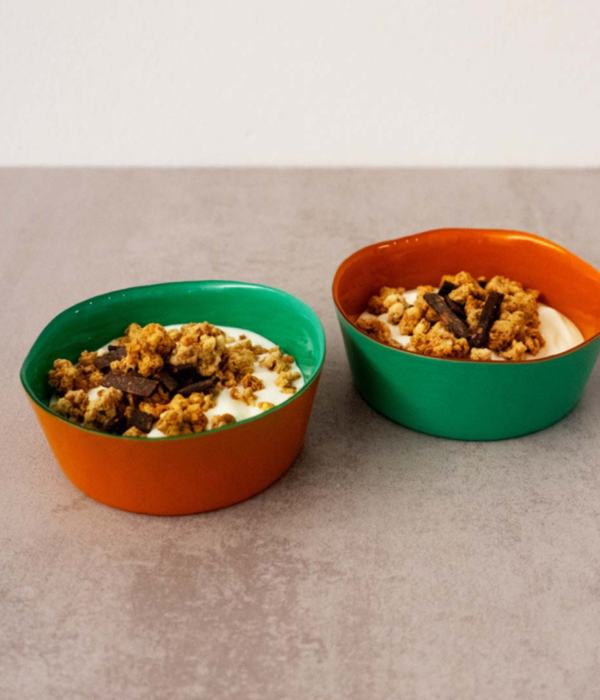 Due coppette vetro, colori Verde e Arancione con dentro yogurt e cereali. Sfondo tavolo grigio.