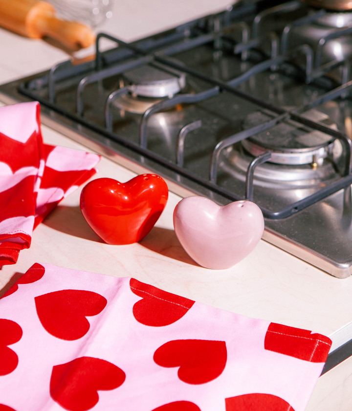 Due cuori in ceramica rosso e rosa su piano cottura. Sfondo gas cucina e strofinacci con cuore.