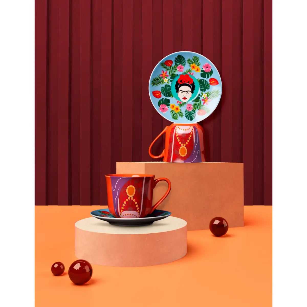 Set tazza e piattino che raffigurano Frida Kahlo. Appoggiati su tavolo arancione. Sfondo rosso