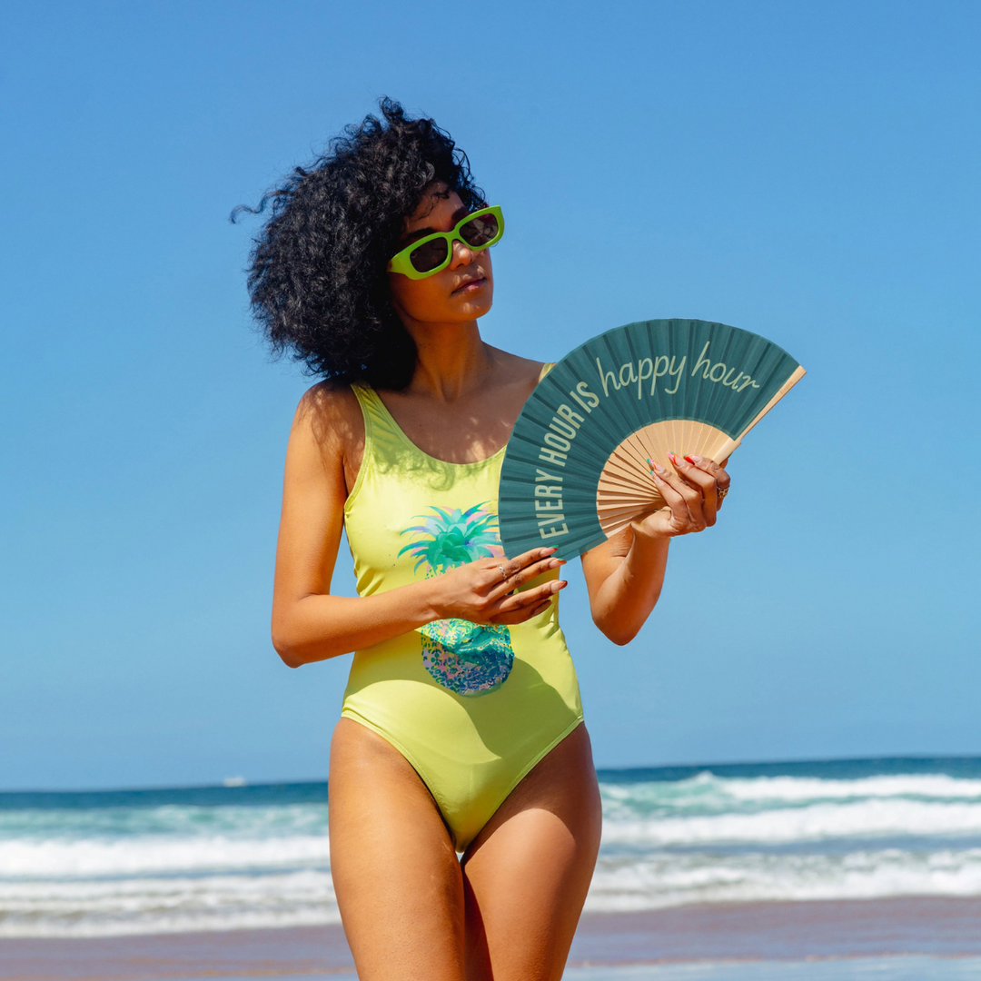 Donna in spiaggia con ventaglio Every hour is happy hour in mano. Costume verde. Sfondo mare 
