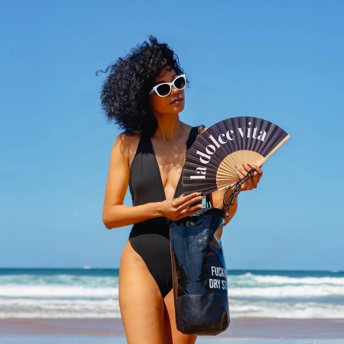 Donna in spiaggia con ventaglio Shake It Off in mano. Costume e borsa da spiaggia nera. Sfondo mare 