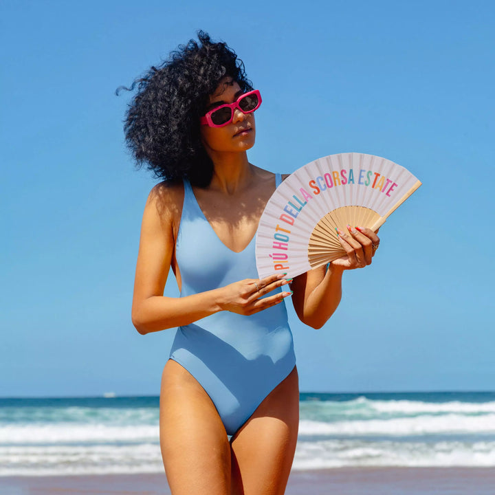 Donna in spiaggia con ventaglio Più Hot della Scorsa Estate in mano. Costume azzurro. Sfondo mare 
