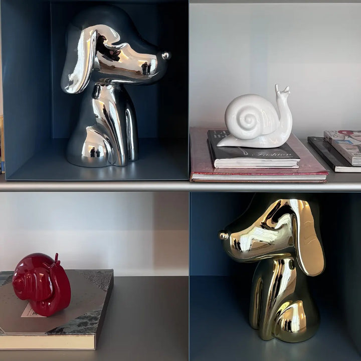 Due statue a forma di cane oro, argento appoggiate dentro una libreria. Libri e lumache di design