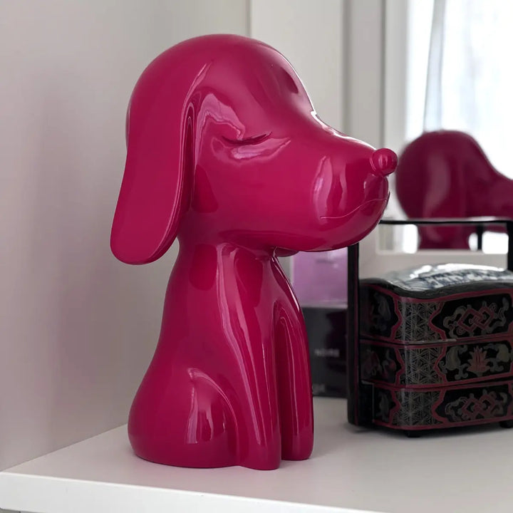 Statua a forma di cane fuxia con occhi naso e bocca appoggiata su mobile bianco. Sfondo specchio
