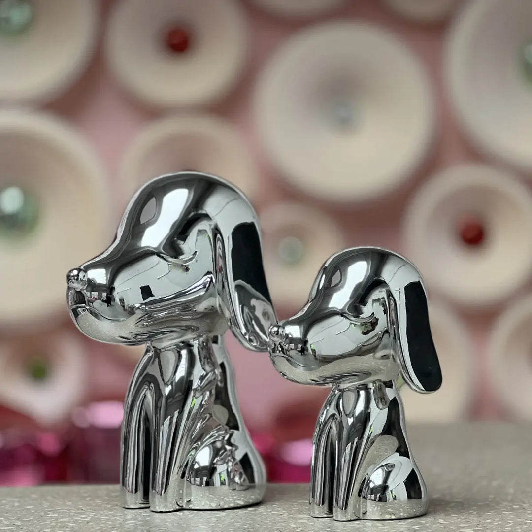 Due statue a forma di cane una grande. e una piccola appoggiate su tavolo grigio. Sfondo rosa