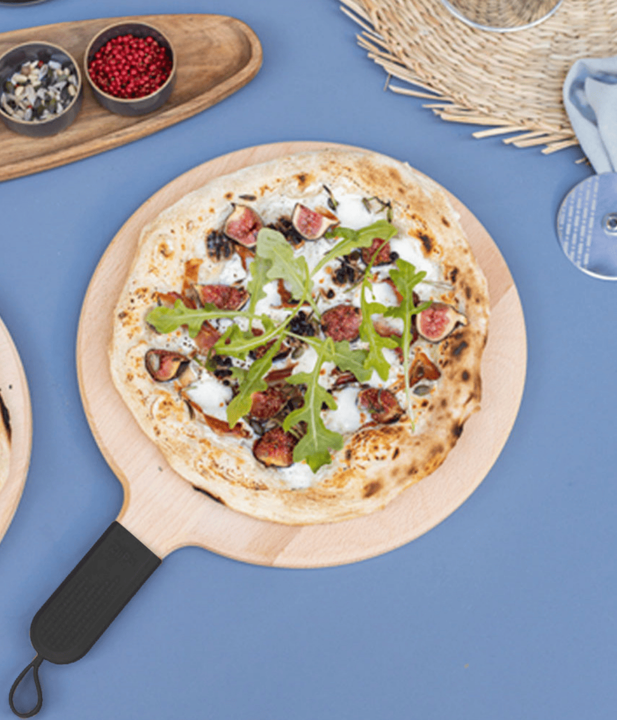 Pizza con rucola e fichi appoggiata su tagliere di legno con manico nero e gancetto. Sfondo tavolo azzurro con accessori tavola.