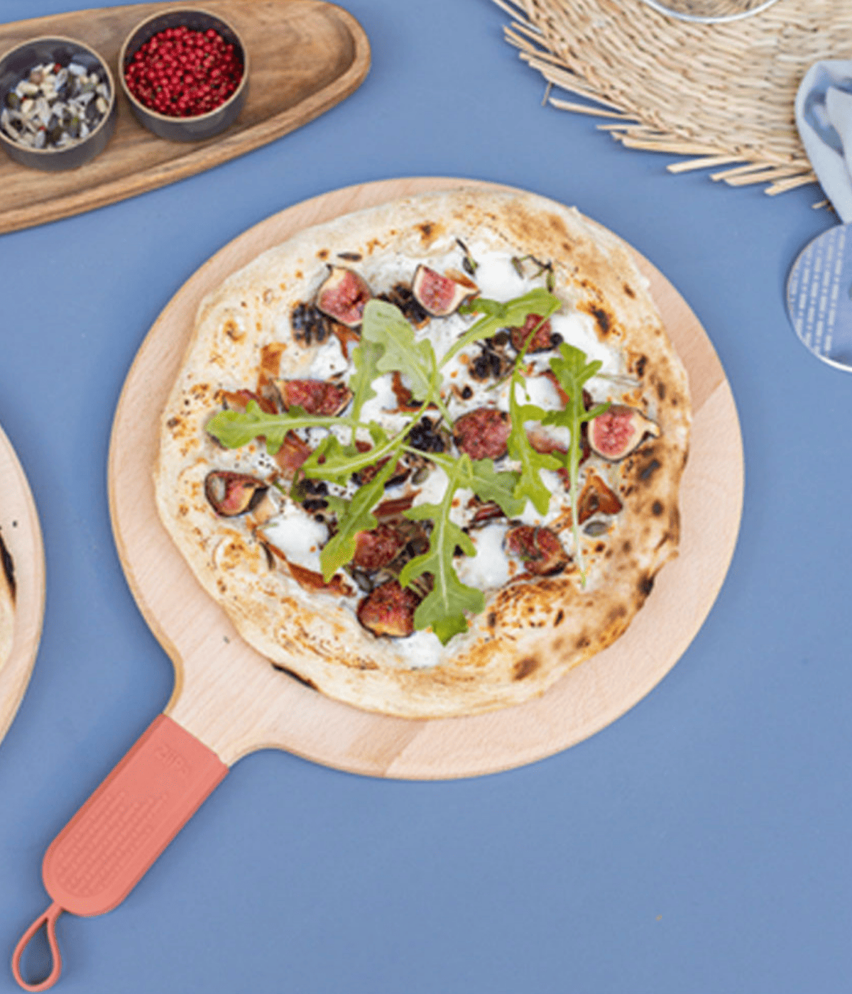 Pizza con rucola e fichi appoggiata su tagliere di legno con manico terracotta e gancetto. Sfondo tavolo azzurro con accessori tavola.