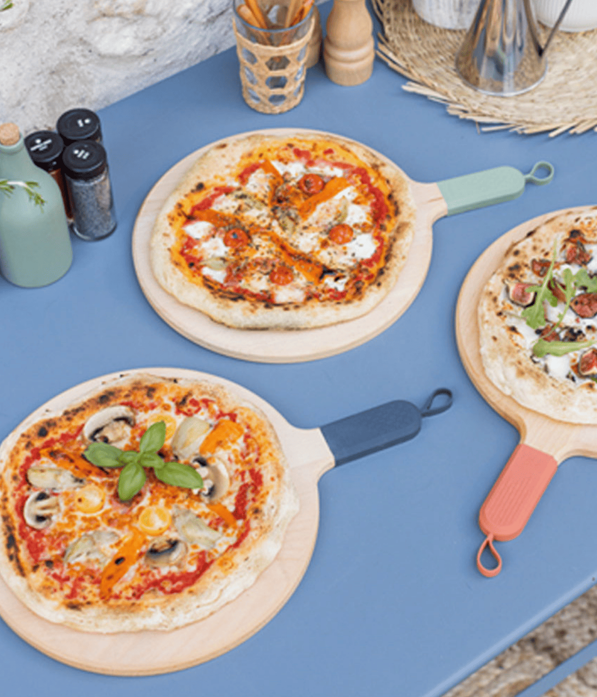Tre pizze appoggiate su taglieri di legno con manico colorato. Sfondo tavolo azzurro con accessori cucina.