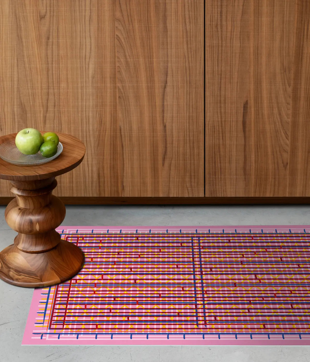 Un tappeto rosa, con righe colorate rosse, gialle, bianche. Tavolino con mele, sfondo mobile marrone