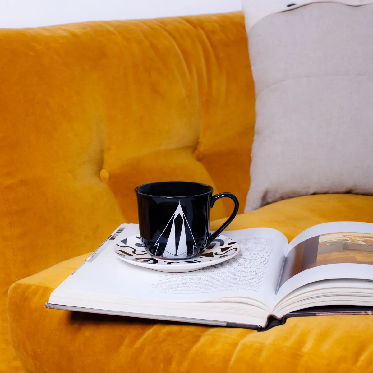 Tazza e piattino nero e bianco, posato su libro aperto, il tutto appoggiato su divano ocra. 