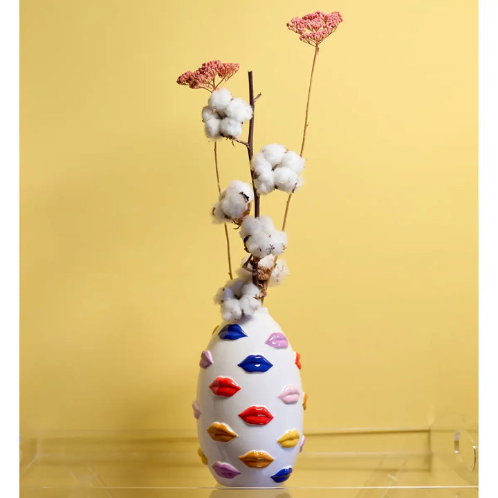 Vaso Mouth con labbra multicolor appoggiato su mensola di vetro con fiori all'interno. Sfondo giallo