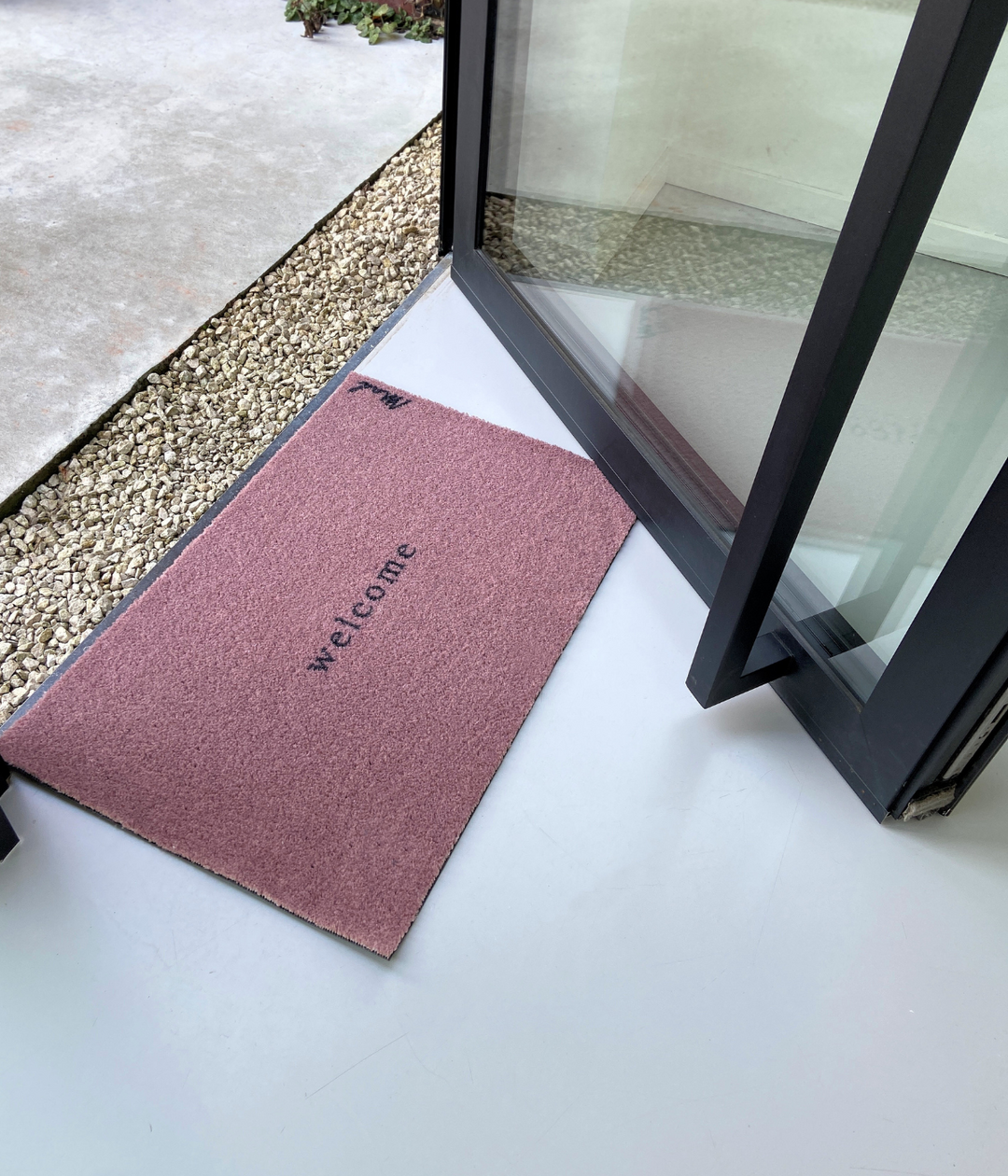 Ingresso con sassi e porta nera con Zerbino Welcome Rosa 50 x 75 cm. Sfondo pavimento bianco