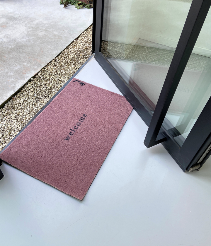Ingresso con sassi e porta nera con Zerbino Welcome Rosa 50 x 75 cm. Sfondo pavimento bianco