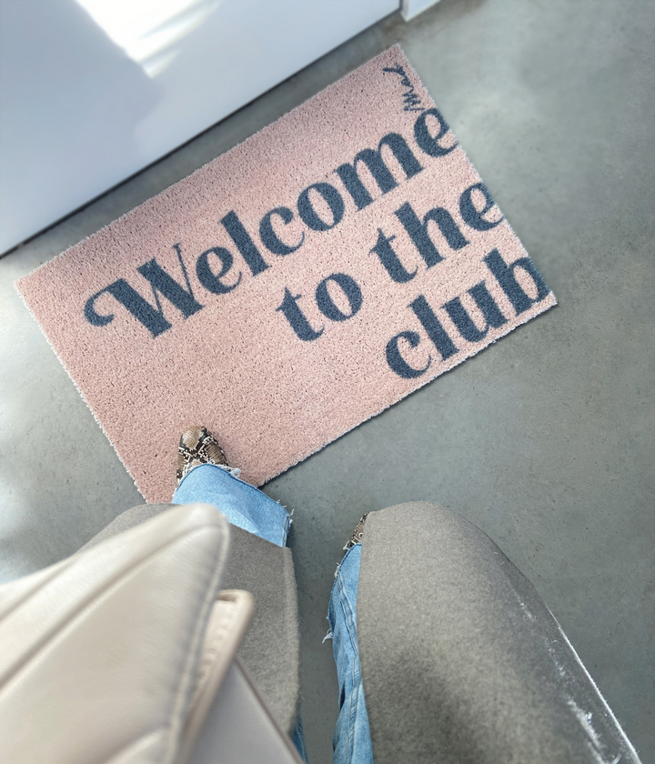 Persona che guarda lo zerbino beige con scritta grigia Welcome to the club. Sfondo pavimento