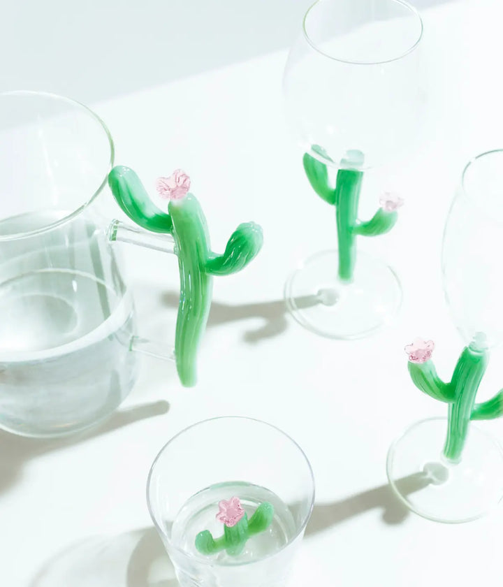 Brocca, calice e bicchiere in vetro con Cactus verde opaco e fiore rosa. Sfondo tavolo bianco
