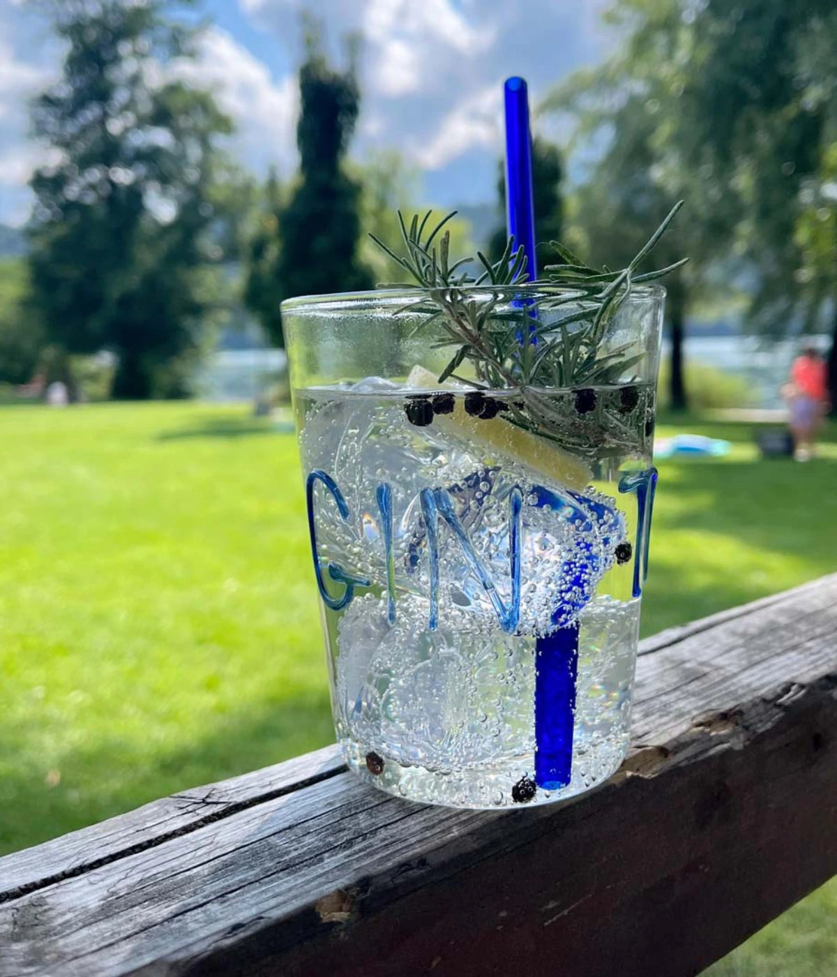 Bicchiere vetro trasparente scritta Gin Tonic e cannuccia blu. Rosmarino e ghiaccio. Sfondo prato