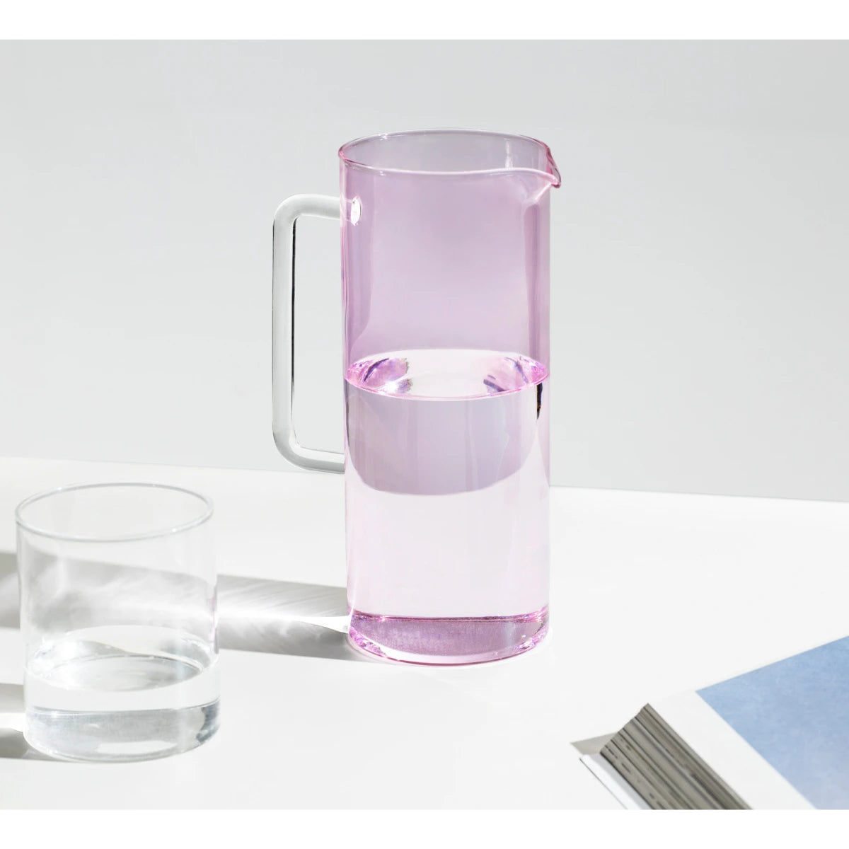 Brocca in vetro rosa con manico in vetro trasparente, bicchiere d'acqua, appoggiati su tavolo bianco