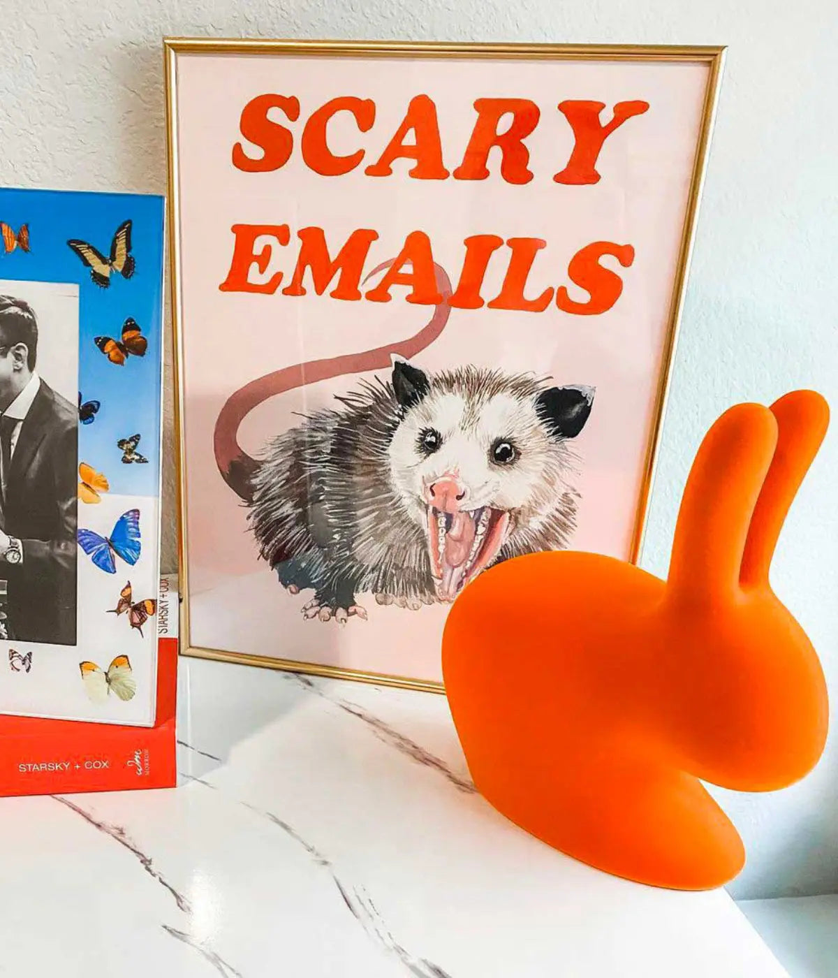 Un coniglio arancione in velluto appoggiatio su tavolo in marmo. Sullo sfondo cornici con disegni.