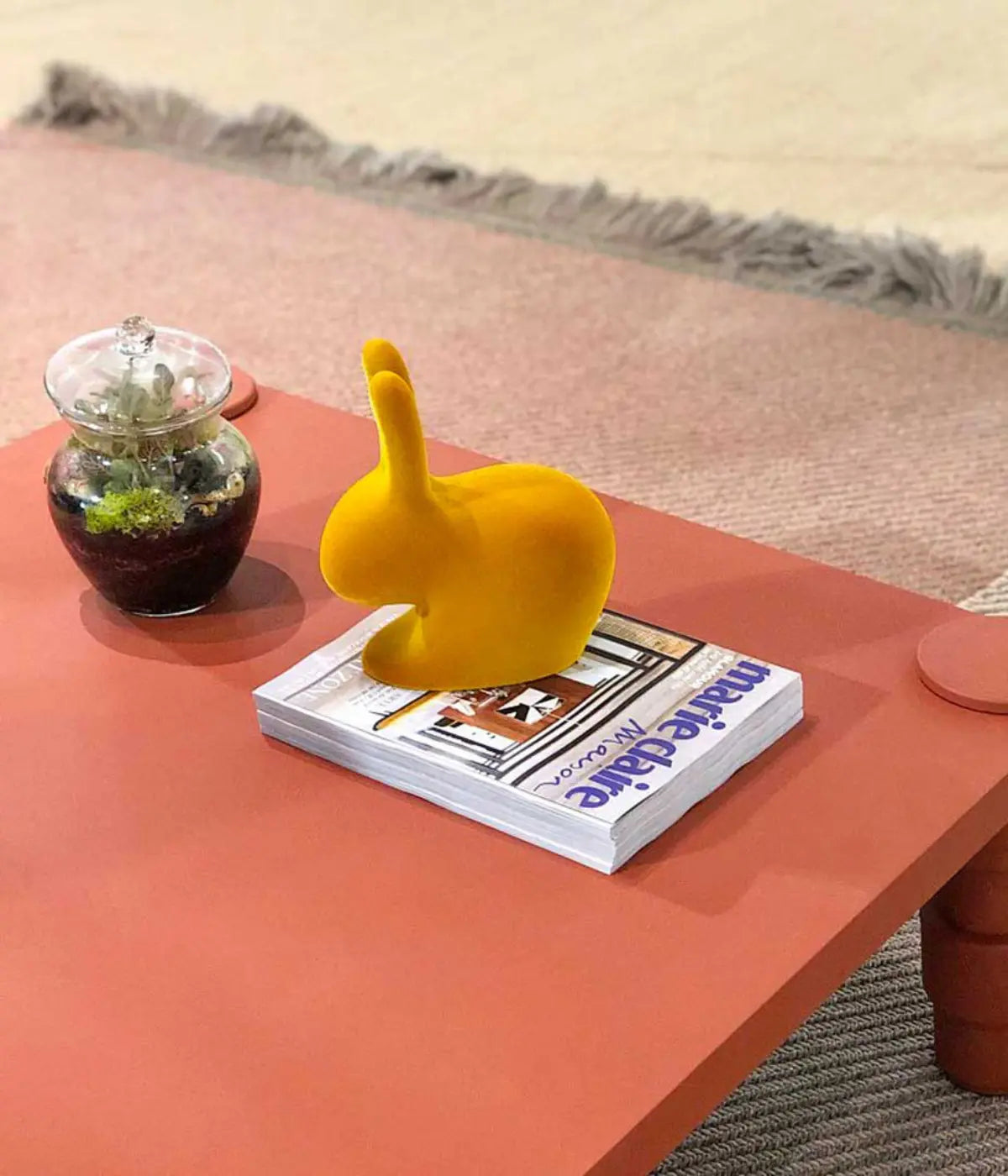 Un coniglio color senape appoggiato su tavolino terracotta e libro. Sfondo tappeto e pianta.