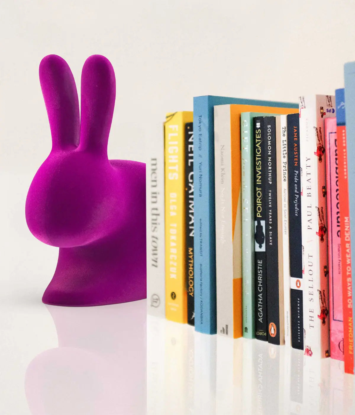 Un coniglio viola in velluto che fa da fermalibri. Libri colorati appoggiati su tavolo bianco.