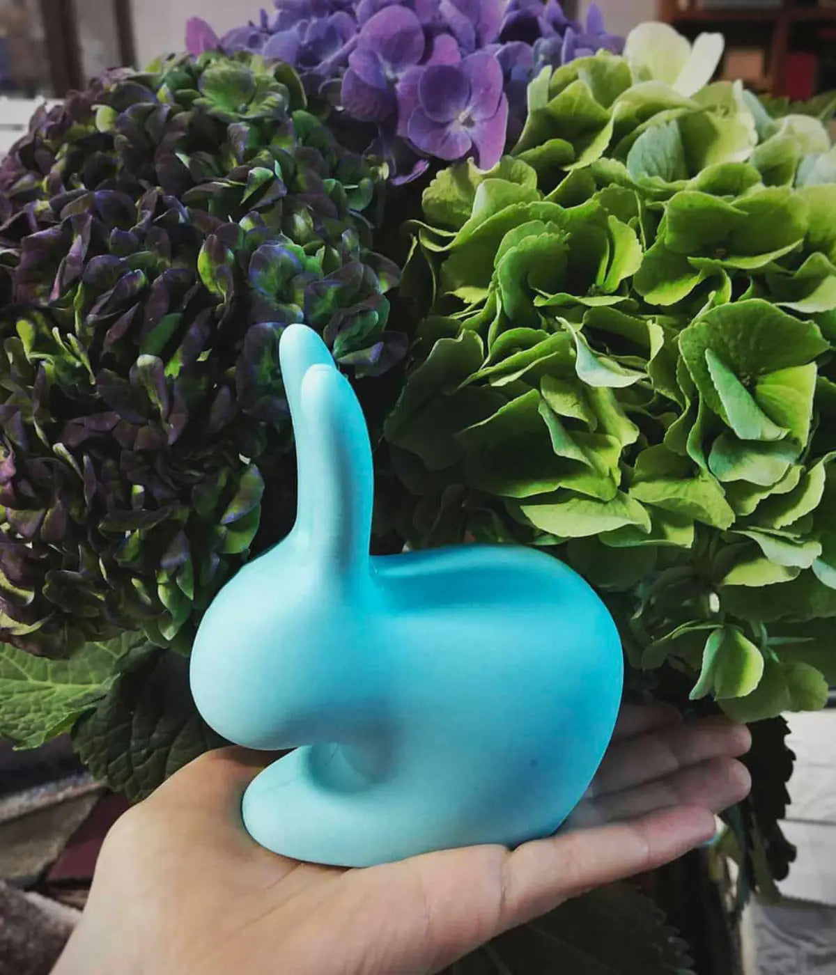 Un fermaporta o fermalibro a forma di coniglio celeste tenuto su una mano. Sfondo ortensie