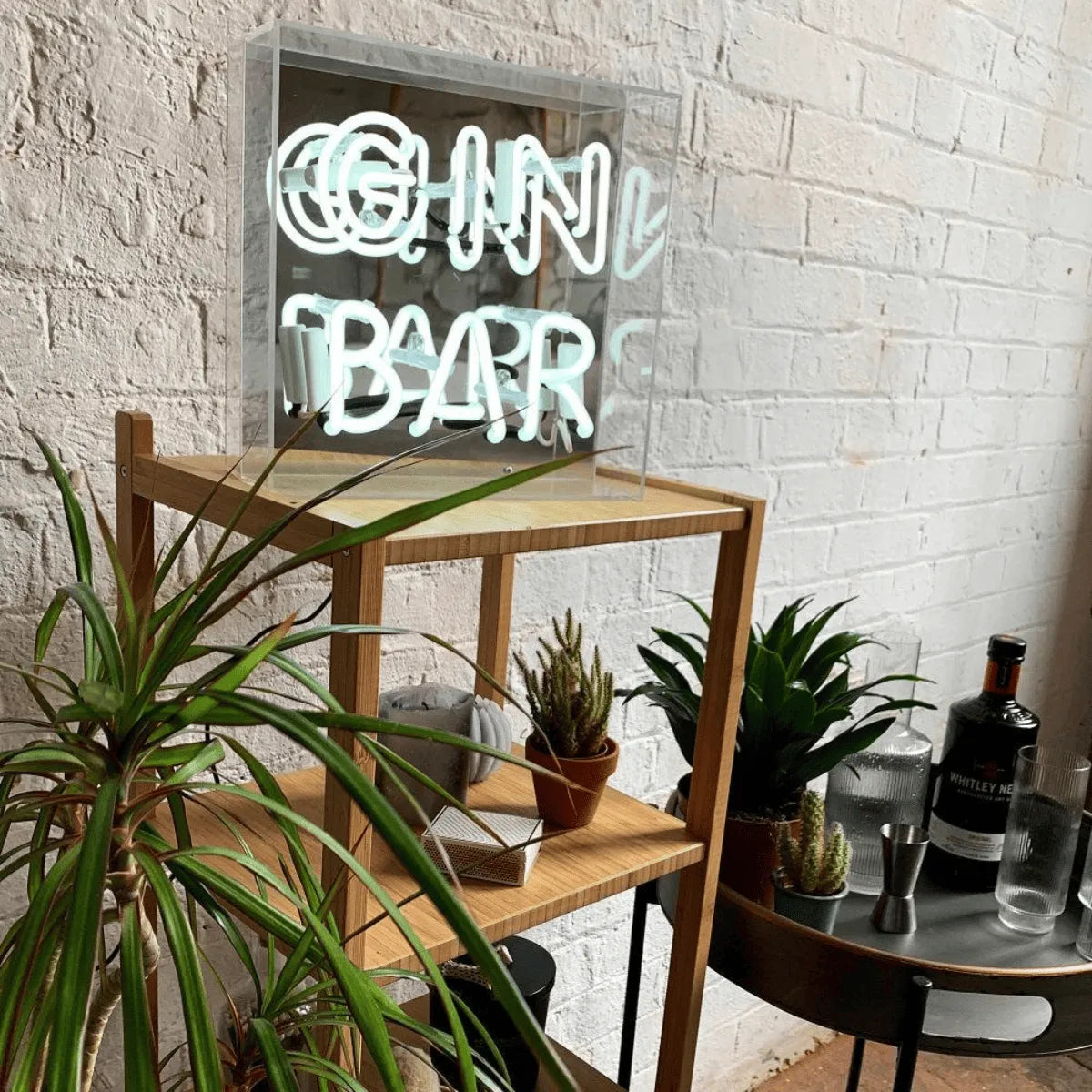 insegna neon illuminata con scritta gin bar su tavolino in legno, circondata da piante e bottiglia.