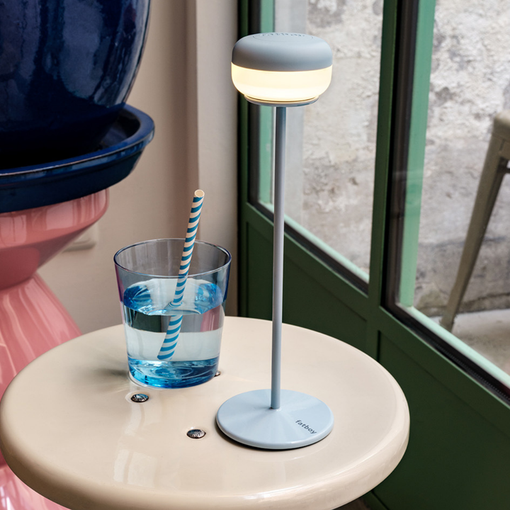 Lampada da tavolo azzurra appoggiata su tavolo beige con vicino bicchiere acqua. Sfondo interno casa