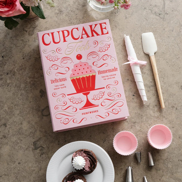 kit cupcake rosa su piano di lavoro con utensili, fiori e piatto con dolcetti.